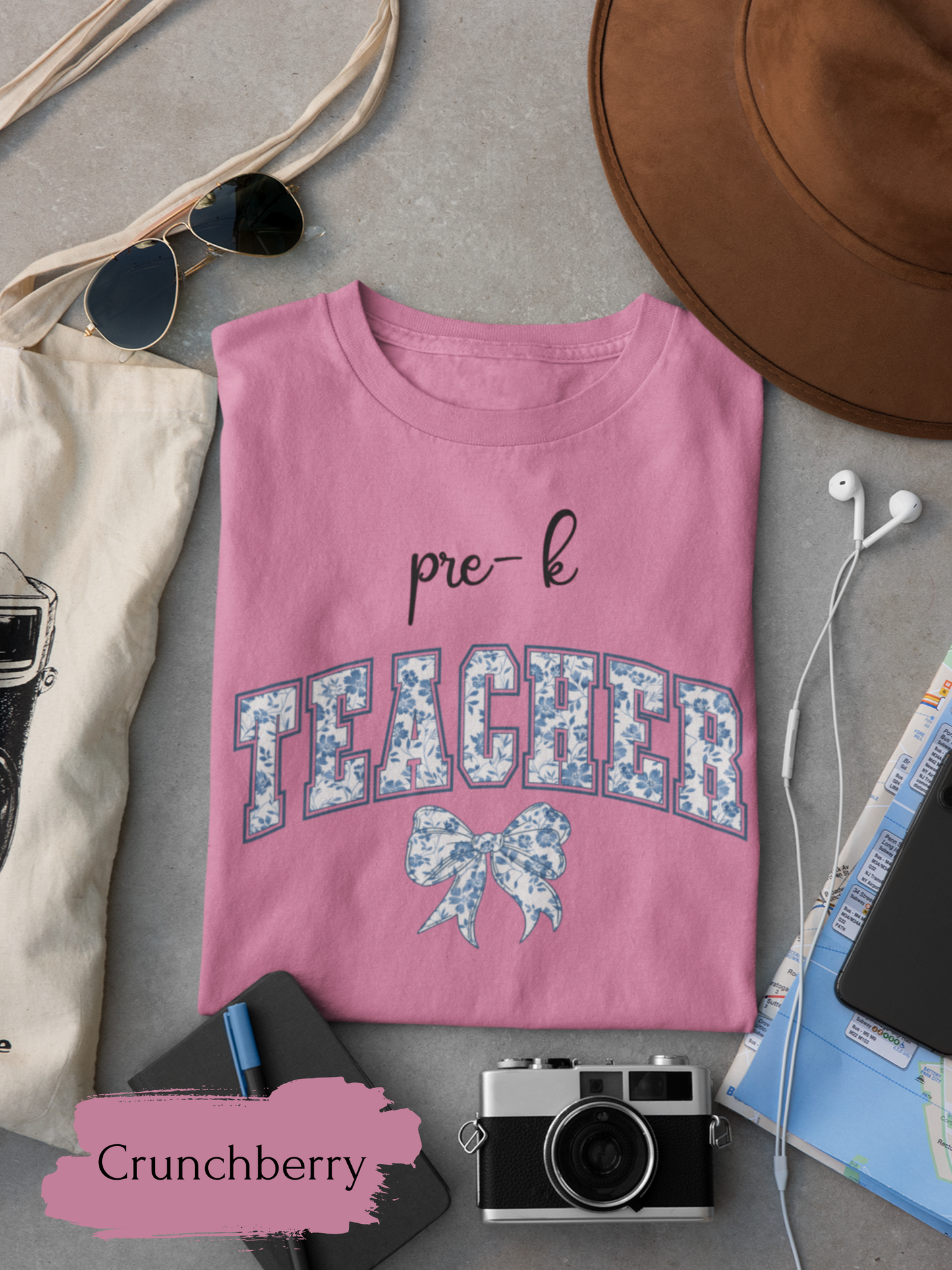 Pink t-shirt with 'pre-k TEACHER' text and bow design, surrounded by accessories on a gray surface.